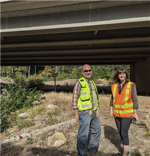 A white man and woman in safety vests stand under a freeway overpass in a forested area